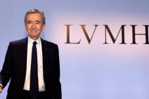 Portrait of Bernard Arnault, the richest person in the world in 2023, smiling in front of the logo of LVMH, the largest luxury-products company in the world.